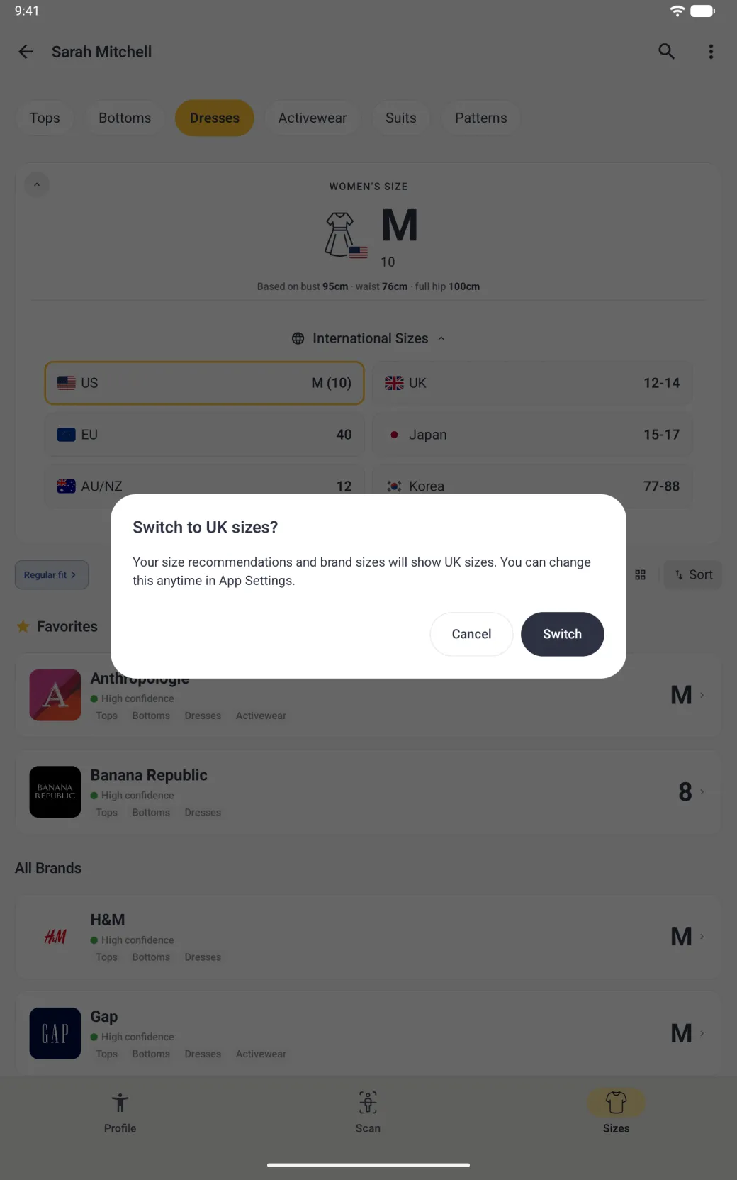 A dialog over the dimmed Sizes tab for Dresses titled Switch to UK sizes? reading Your size recommendations and brand sizes will show UK sizes. You can change this anytime in App Settings., with an outlined Cancel button and a filled navy Switch button; the International Sizes tiles show behind it with US outlined in gold.