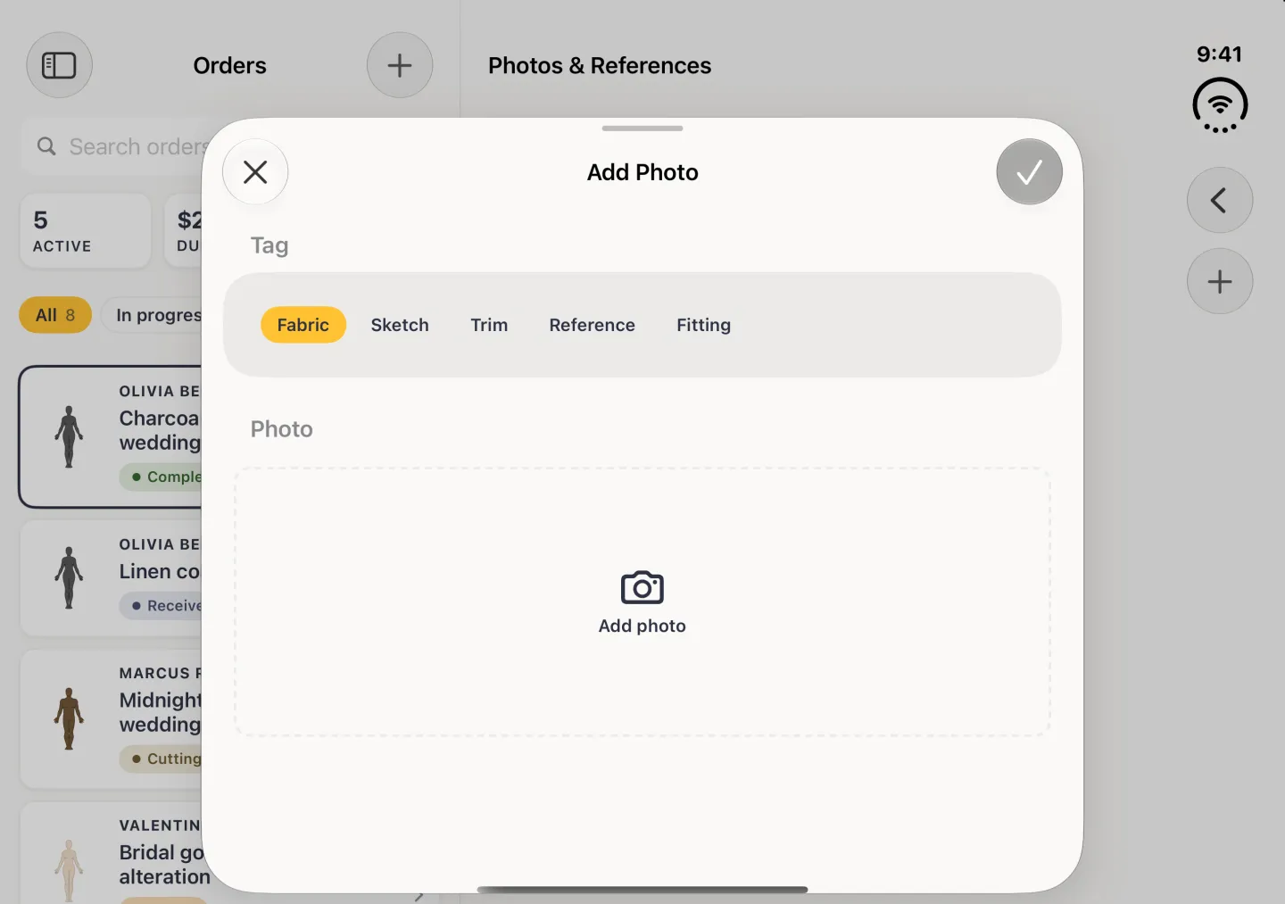 Add Photo sheet centred over the dimmed Orders column and Photos and References pane in landscape, with a grab handle, a close button at the top left and a dimmed round tick at the top right, tag chips reading Fabric, Sketch, Trim, Reference and Fitting with Fabric selected in gold, and an empty dashed Add photo area with a camera icon.